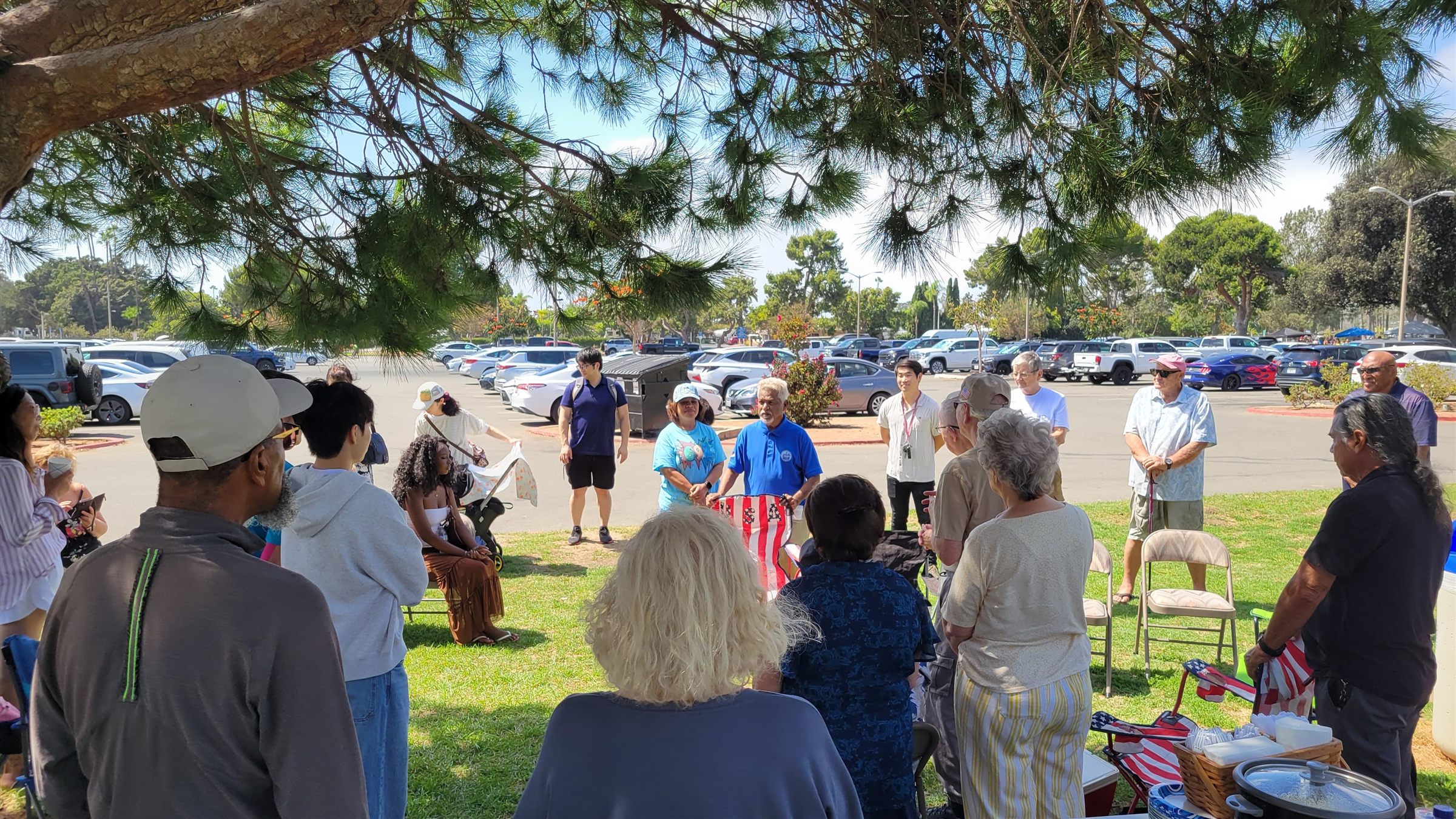 Church members gathered under a pine tree at a park picnic with tables of food