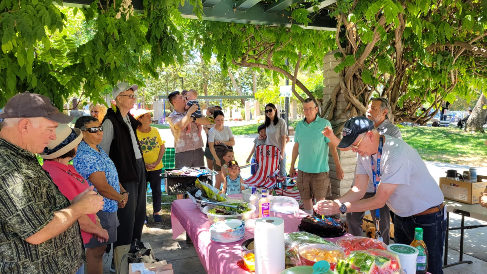 Church families gathered around a picnic table of food under a wisteria arbor
